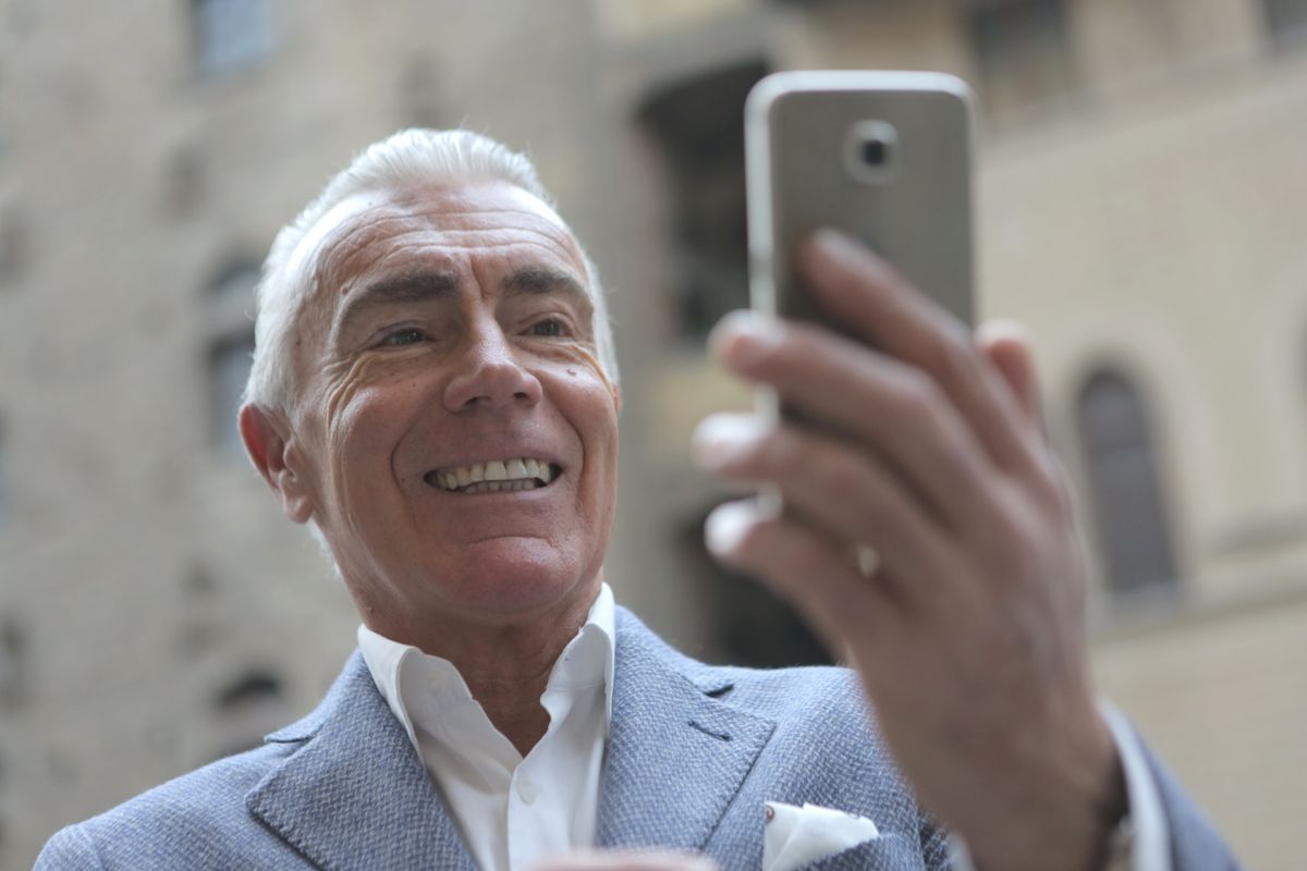A smiling elderly gentleman looking directly at the camera while holding his smartphone